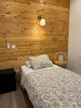 Cama individual con lámpara de pared encendida y mininevera junto a la pared de madera